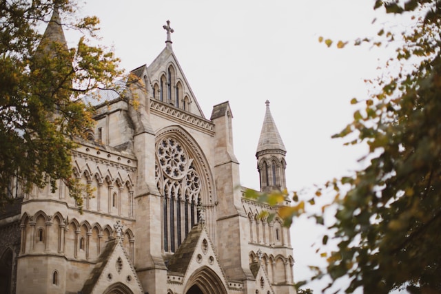 Gothic stone church in Hertfordshire with arched windows, carvings, and rose window, trees edging the scene—think local impact.